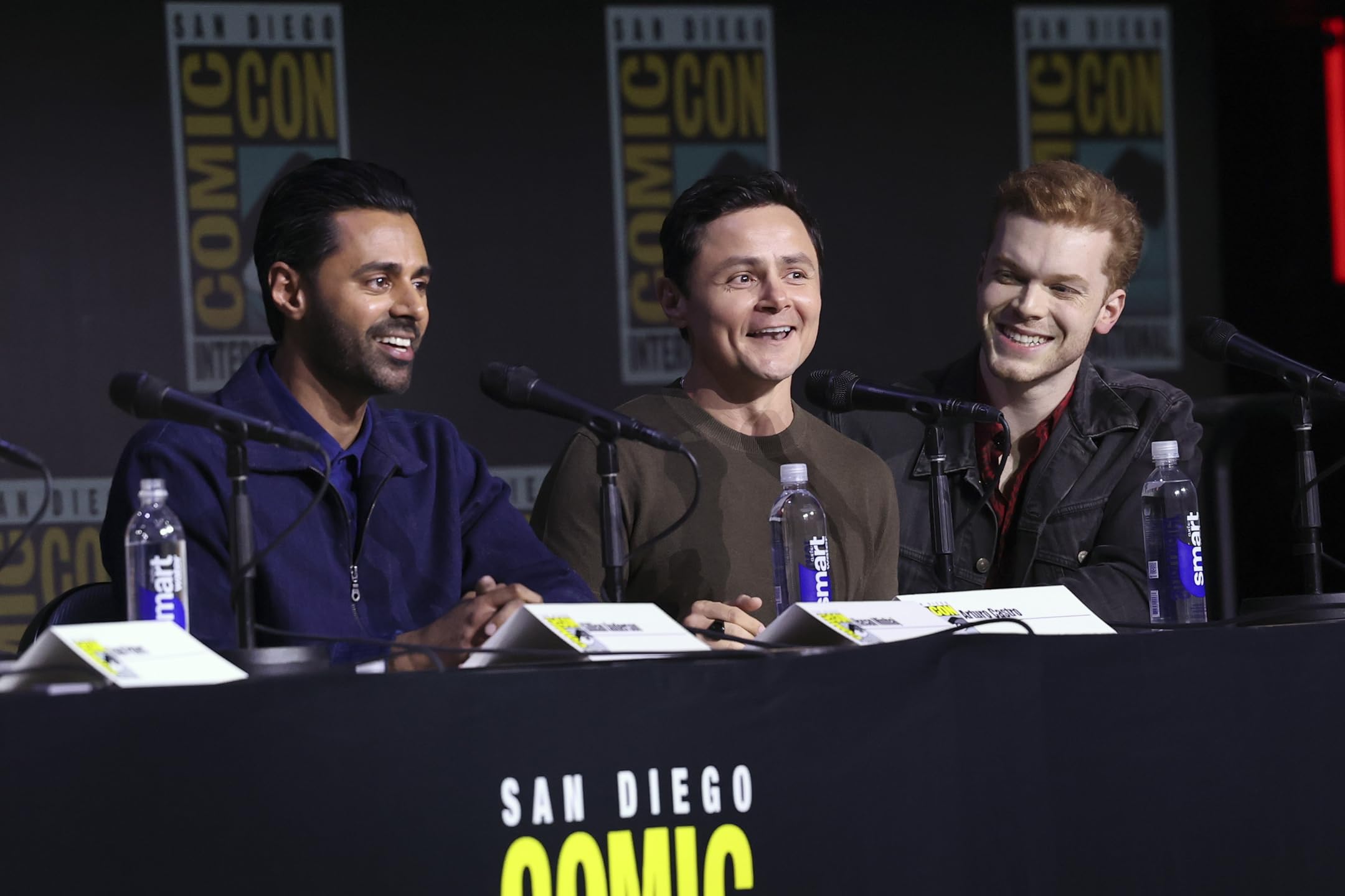 Evan Peters, Gillian Anderson, and Hasan Minhaj speaking on the Tron: Ares Comic-Con panel, discussing the upcoming film with fans and press.
