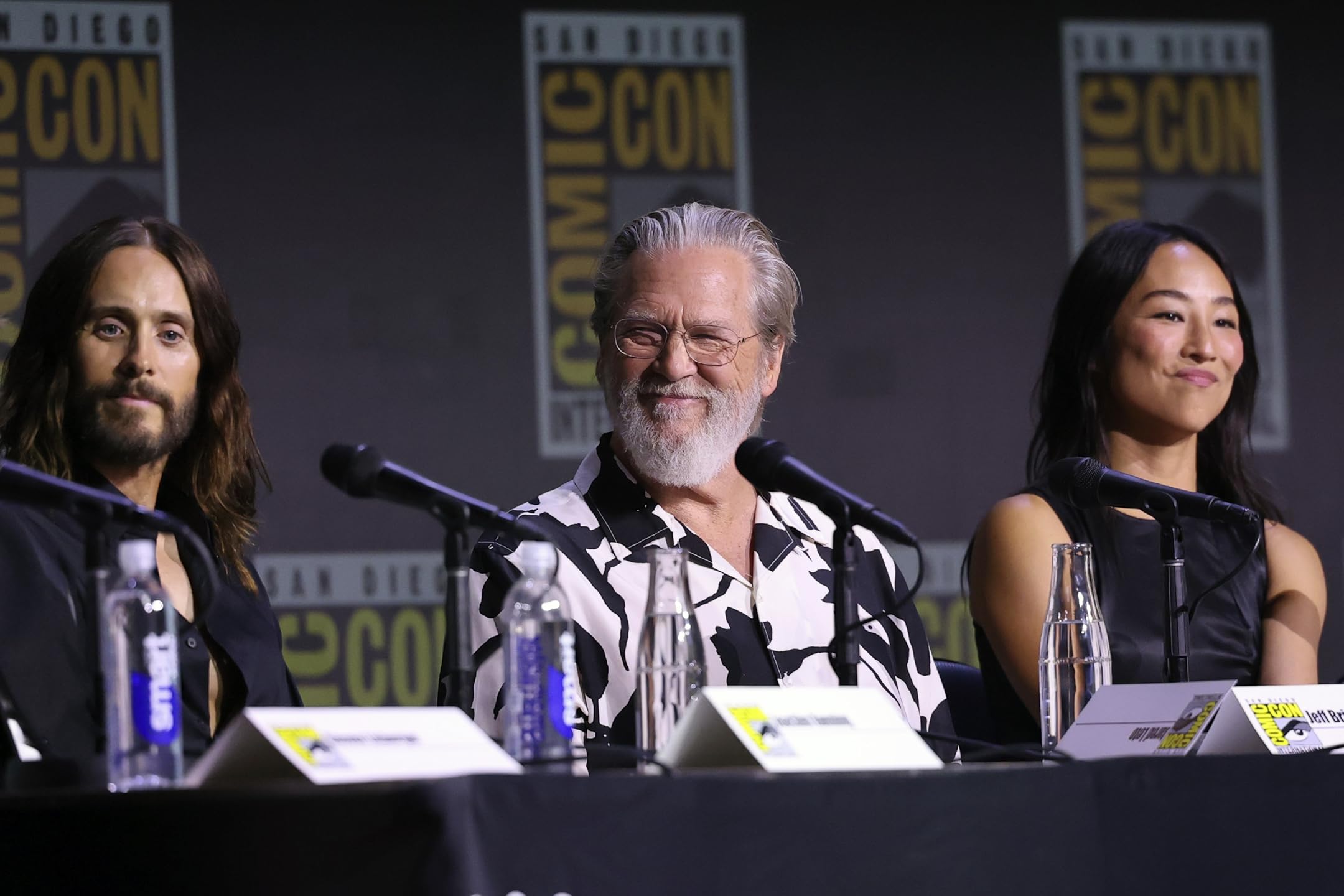 Evan Peters, Gillian Anderson, and Hasan Minhaj speaking on the Tron: Ares Comic-Con panel, discussing the upcoming film with fans and press.
